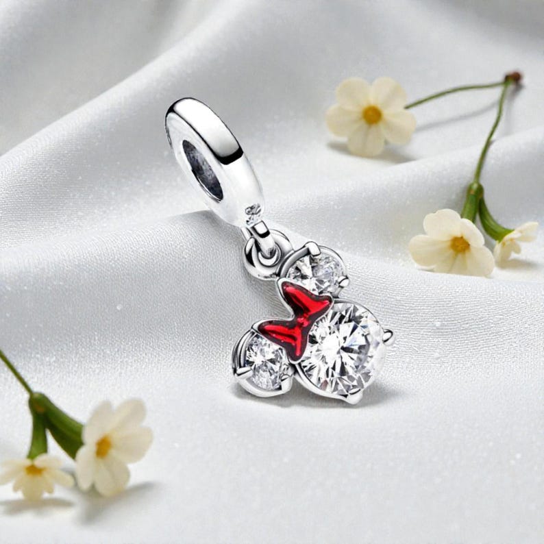 May include: A silver charm with a Minnie Mouse design. The charm features three clear, sparkling cubic zirconia stones and a red enamel bow. The charm is suspended from a silver loop, and is set against a white, silky background.