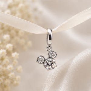 May include: A silver-toned metal charm pendant featuring three round, clear, faceted stones arranged in a cluster. The stones are held in place by small metal prongs, and the charm is suspended from a polished silver-toned metal jump ring.