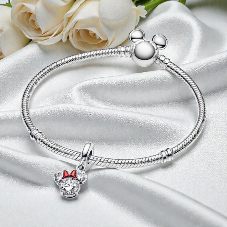 May include: A silver bracelet with a Mickey Mouse head clasp and a Minnie Mouse charm with a red bow. The bracelet is on a white satin surface with white roses in the background.
