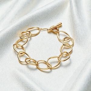 May include: A gold-colored bracelet with oval links and a toggle clasp. The bracelet is displayed on a white, silky surface, creating a contrast that highlights the jewelry's design and color. The links are connected in a chain.