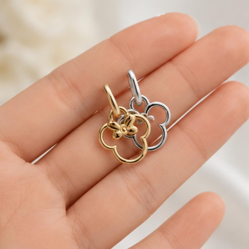 Puede incluir: Dos dijes de metal, uno de tono dorado y otro de tono plateado, cada uno con forma de flor de cuatro p&eacute;talos con un peque&ntilde;o detalle de lazo en la parte superior. Cada dije cuenta con una anilla circular en la parte superior para su fijaci&oacute;n.