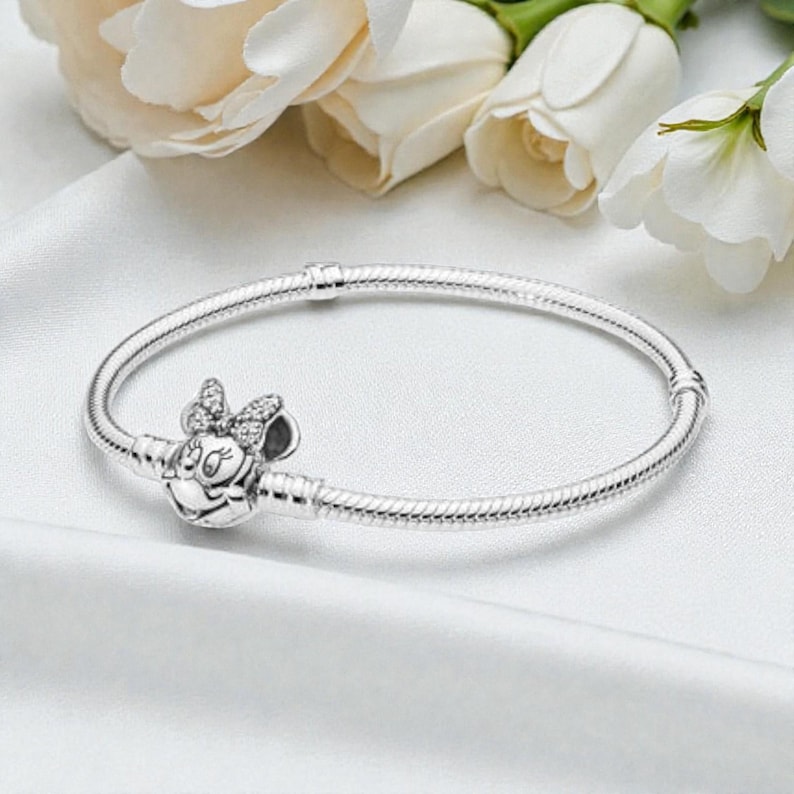 May include: A silver snake chain bracelet featuring a clasp shaped like a cartoon mouse head with a bow. The bow is embellished with small clear stones, and the bracelet includes two fixed silver stations along the circular band structure.
