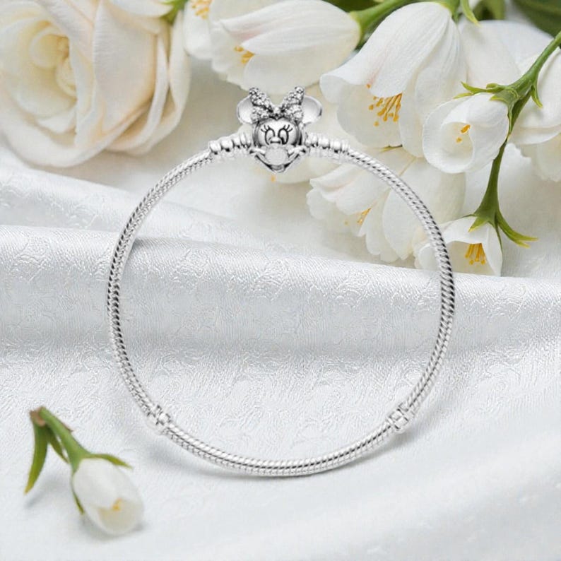 May include: A silver snake chain bracelet featuring a clasp shaped like a cartoon mouse head with a bow. The bow is embellished with small clear stones. The bracelet has a smooth, rounded metallic texture and a circular shape.