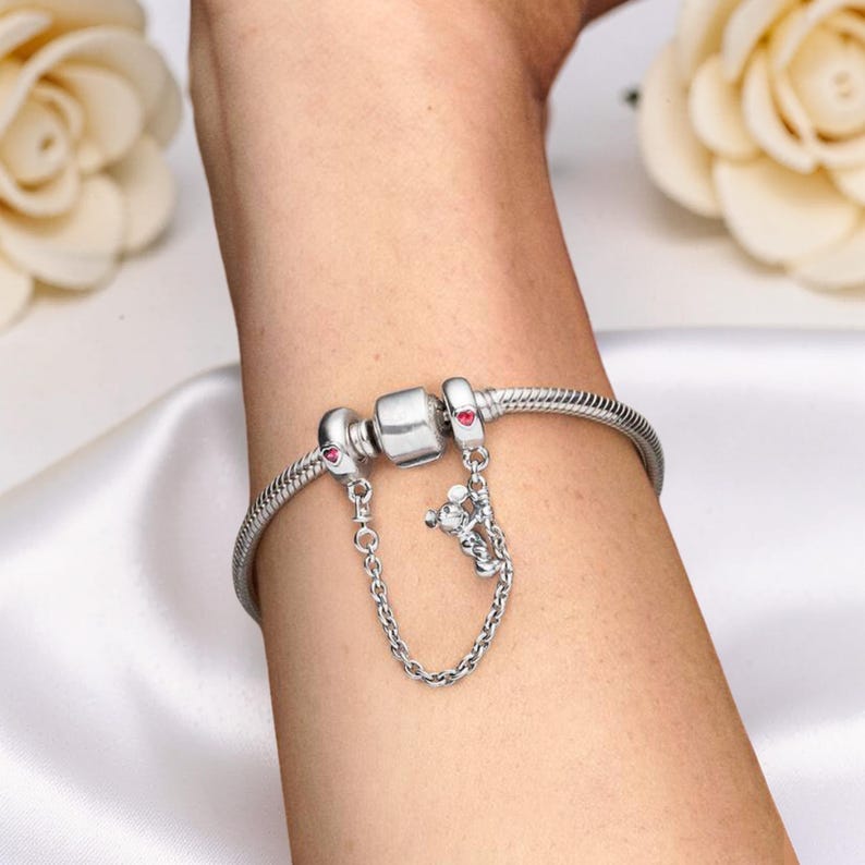 May include: A silver bracelet with a snake chain design. The bracelet features a safety chain with a small charm and red accents. The bracelet is secured with a clasp. The bracelet is shown on a person's wrist.