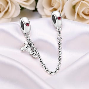May include: A silver charm bracelet safety chain with two barrel-shaped ends, each featuring a red heart-shaped accent. A small Mickey Mouse charm hangs from one end. The chain is made of small, interconnected links.
