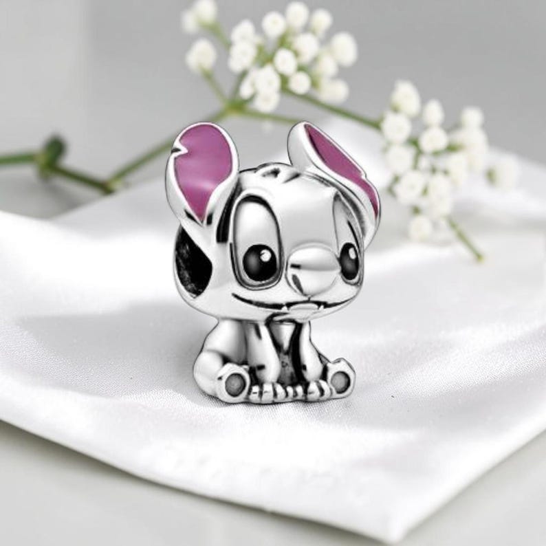 May include: A silver charm bead shaped like the character Stitch, featuring large black eyes, a small nose, and prominent ears with pink enamel detailing. The metallic surface is polished and reflective, showcasing the character in a seated position.