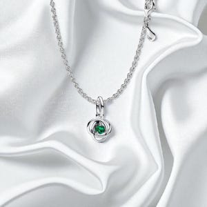 May include: A silver necklace with a pendant featuring a green gemstone set in a stylized knot design. The chain is delicate, and the pendant is centered. The necklace is displayed on a white, silky fabric.