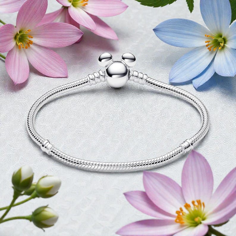 Pu&ograve; includere: Un bracciale d'argento con un ciondolo a forma di Mickey Mouse. Il bracciale ha un design a catena a serpente ed &egrave; circondato da fiori rosa e blu. Il ciondolo presenta una perla rotonda con due perle pi&ugrave; piccole sopra.