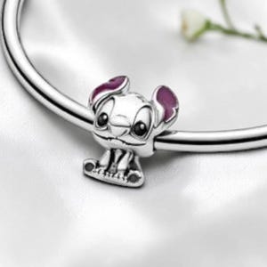 May include: A silver charm bead shaped like the character Stitch, featuring large black eyes and purple enamel accents on the inner ears, threaded onto a polished silver bracelet band. The charm displays a detailed facial expression.