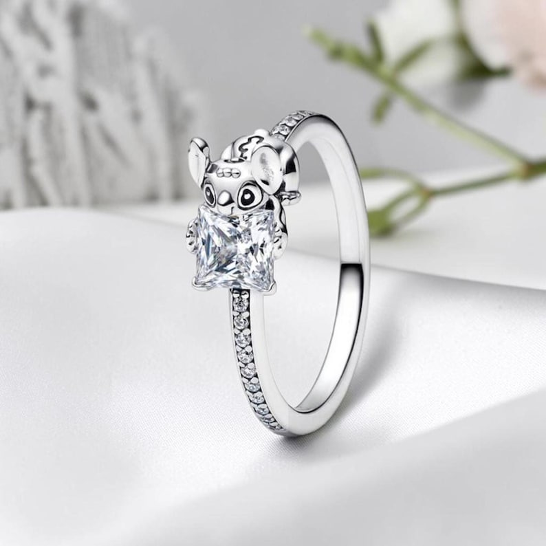 May include: A silver-toned ring featuring a character figure holding a square-cut clear gemstone. The band is adorned with a row of small, round, clear stones along the shank, providing a sparkling finish to the metallic design.