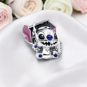 May include: A silver charm featuring a cartoon character wearing a graduation cap and holding a scroll. The charm has black, blue, and pink accents. The background includes white fabric and blurred flowers.