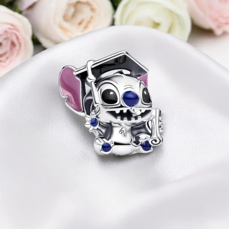 May include: A silver charm featuring a cartoon character wearing a graduation cap and holding a scroll. The charm has black, blue, and pink accents. The background includes white fabric and blurred flowers.