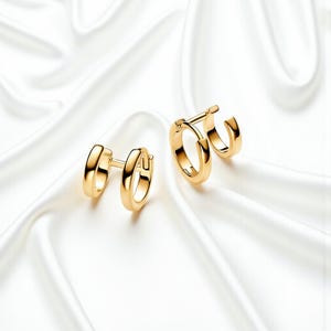May include: Two gold-colored hoop earrings. One pair features a double-hoop design, while the other has a single hoop with a smaller, attached hoop. The earrings are displayed on a white, silky surface.
