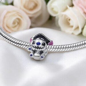 May include: A silver charm featuring a cartoon character wearing a graduation cap. The charm has blue eyes and purple accents. It is attached to a silver bracelet with a textured design. The background includes soft focus white and pink flowers.