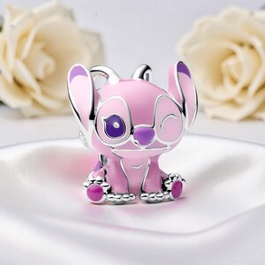 Peut inclure: Un charm rose et argent&eacute; repr&eacute;sentant un personnage de dessin anim&eacute; avec de grandes oreilles et une expression qui cligne de l'&oelig;il. Le charm a des accents violets sur les oreilles, les yeux et les pieds. Le charm est pos&eacute; sur un fond blanc avec des fleurs floues.