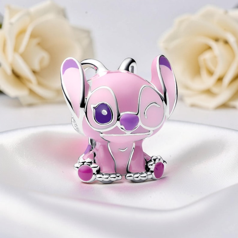 Peut inclure: Un charm rose et argent&eacute; repr&eacute;sentant un personnage de dessin anim&eacute; avec de grandes oreilles et une expression qui cligne de l'&oelig;il. Le charm a des accents violets sur les oreilles, les yeux et les pieds. Le charm est pos&eacute; sur un fond blanc avec des fleurs floues.