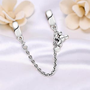 May include: A silver charm bracelet safety chain with a Mickey Mouse charm. The chain has two clasps with a curved black design and a small chain connecting them. The Mickey Mouse charm is detailed and adds a playful touch.