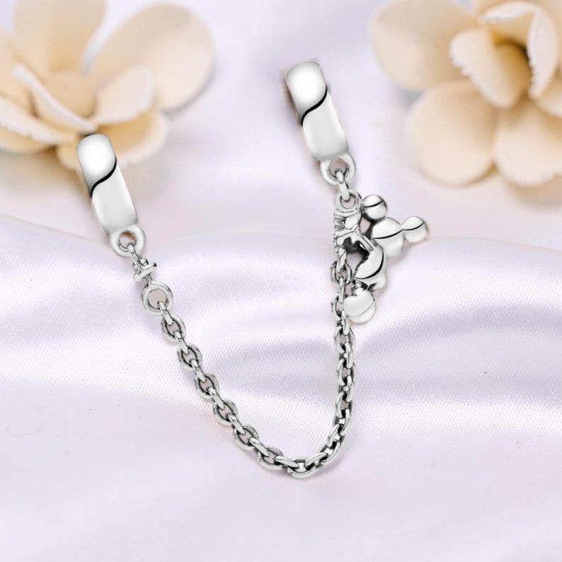 May include: A silver charm bracelet safety chain with a Mickey Mouse charm. The chain has two clasps with a curved black design and a small chain connecting them. The Mickey Mouse charm is detailed and adds a playful touch.