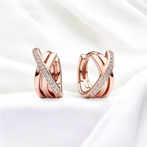 Könnte beinhalten: Ein Paar roségoldene Creolen. Jeder Ohrring hat ein kreuzförmiges Design, wobei ein Band mit kleinen, funkelnden Kristallen besetzt ist. Die Ohrringe werden auf einer weißen, strukturierten Oberfläche präsentiert.