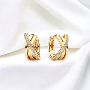 以下が含まれることがあります： 金色のフープピアス2つ。交差したデザインで、各ピアスは片側に小さな透明な宝石が並んでいます。ピアスは白く、質感のある背景に表示されています。