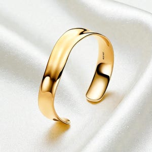 May include: A gold-colored cuff bracelet with a smooth, curved design. The open-ended bracelet has a polished finish, reflecting light. The inside of the bracelet is marked with the text "ALE MET". The jewelry is displayed on a white, silky surface.
