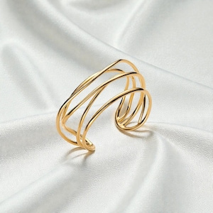 Peut inclure: Un bracelet manchette doré au design moderne. Le bracelet est composé de plusieurs bandes courbes et parallèles, créant un aspect ouvert et aéré. Le métal est poli, reflétant la lumière et ajoutant une touche d'élégance. Le bijou est posé sur un fond blanc et soyeux.