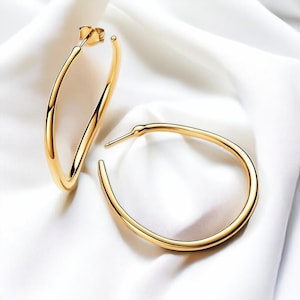 Op de afbeelding: Twee goudkleurige oorringen. De oorbellen hebben een gladde, gepolijste afwerking en een klassiek, minimalistisch ontwerp. Ze zijn geplaatst tegen een witte, zijdeachtige achtergrond, die hun elegante vorm en reflecterende oppervlak benadrukt. Een tijdloos sieraad.