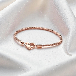 Puede incluir: Pulsera de oro rosa con cierre en forma de corazón. La pulsera tiene un diseño tejido y texturizado. El cierre tiene forma de corazón. La pulsera se muestra sobre una superficie blanca y sedosa.