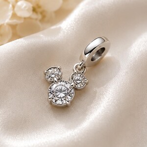 May include: A silver-toned metal charm featuring three round, clear, faceted stones arranged in a cluster resembling a mouse head silhouette. The charm includes a polished circular bail at the top for attachment to a bracelet or necklace chain.