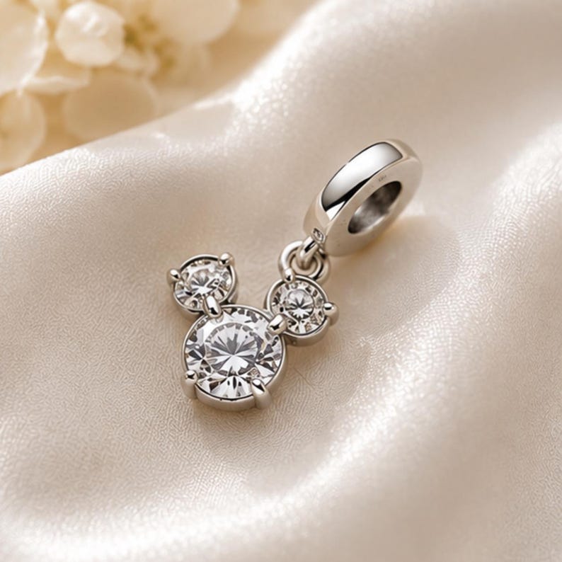 May include: A silver-toned metal charm featuring three round, clear, faceted stones arranged in a cluster resembling a mouse head silhouette. The charm includes a polished circular bail at the top for attachment to a bracelet or necklace chain.