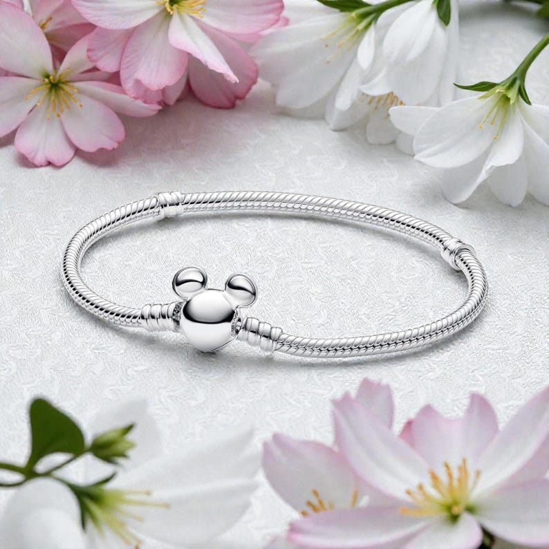 Pu&ograve; includere: Un braccialetto d'argento con chiusura a forma di testa di Topolino. Il braccialetto ha un design a catena a serpente. Fiori rosa e bianchi adornano lo sfondo.