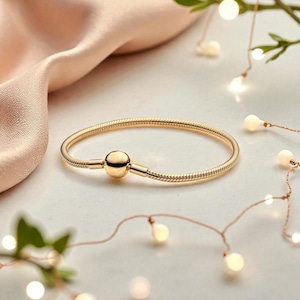 May include: A gold-colored bracelet with a snake chain design. The bracelet features a round clasp in the center. The background includes a soft pink fabric, string lights, and greenery, creating a festive and elegant aesthetic.