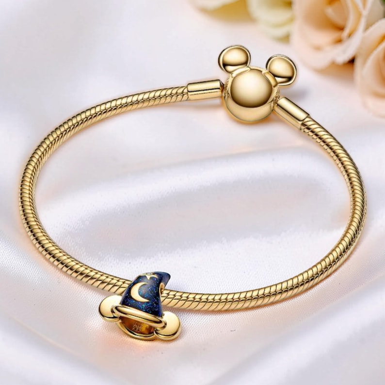 May include: A gold-colored bracelet with a Mickey Mouse-shaped clasp. The bracelet features a charm shaped like a wizard's hat, with a dark blue top, gold trim, and a crescent moon and star design. The bracelet is on a white satin surface.