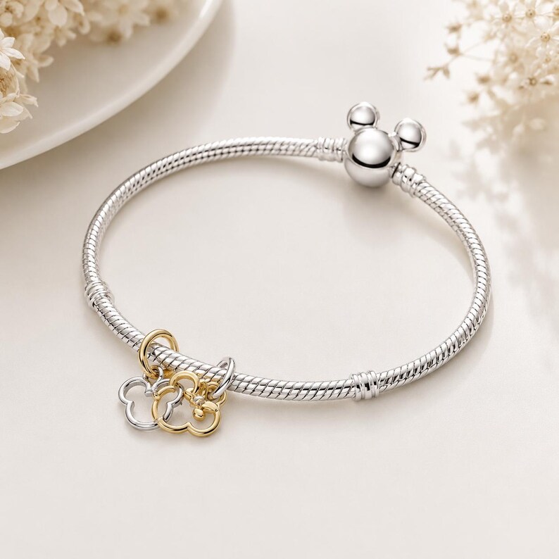 Puede incluir: Una pulsera de cadena de serpiente plateada con un cierre redondo en forma de cabeza de rat&oacute;n. La pulsera incluye dos dijes colgantes, uno plateado y otro dorado, ambos con forma de cabeza de rat&oacute;n con una peque&ntilde;a piedra clara en el dorado.