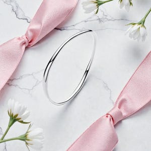 May include: A silver bangle bracelet with a smooth, curved design. The bracelet is displayed on a white marble surface, accented by pink ribbons and white flowers. The bracelet's polished surface reflects light, creating a bright and elegant look.