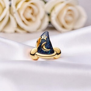 May include: A gold-toned wizard hat-shaped ring with a dark blue, glittery surface. The hat features a crescent moon and star design, with a small diamond accent. The ring is set against a white satin background.