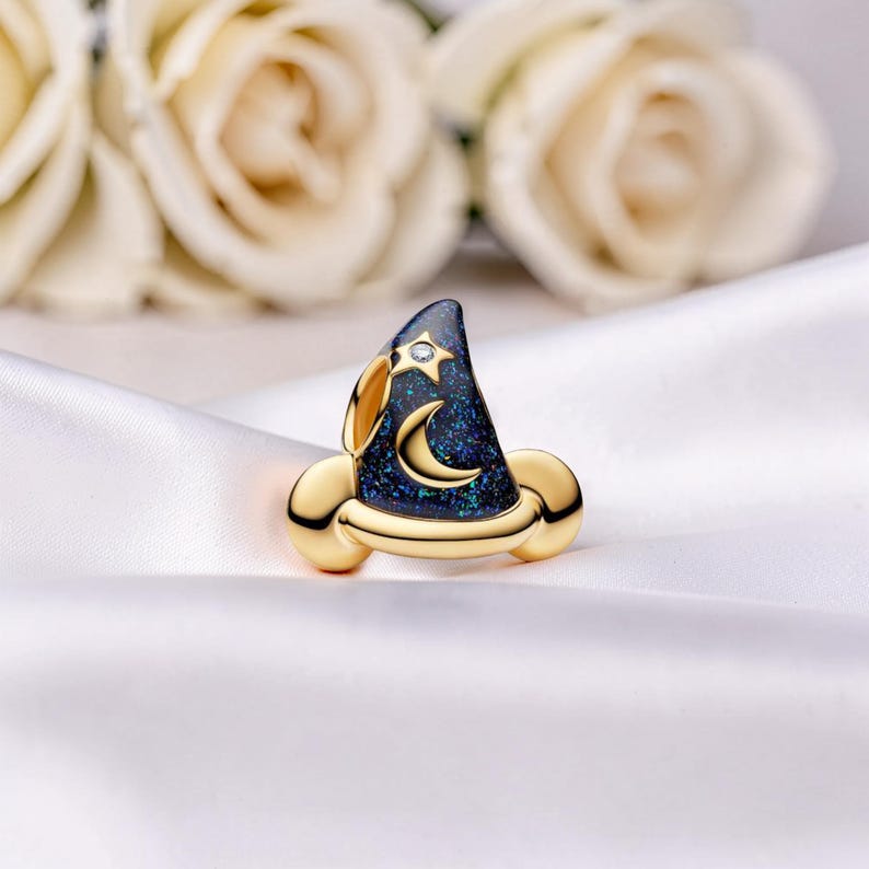 May include: A gold-toned wizard hat-shaped ring with a dark blue, glittery surface. The hat features a crescent moon and star design, with a small diamond accent. The ring is set against a white satin background.
