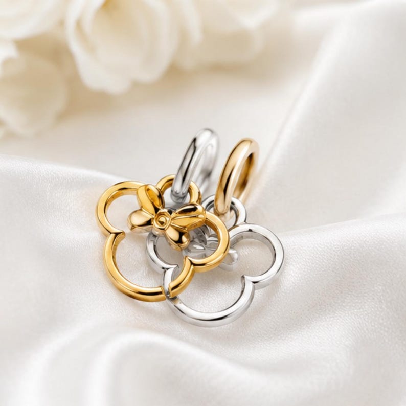 Puede incluir: Dos dijes entrelazados en forma de tr&eacute;bol, uno en oro pulido y otro en plata pulida, cada uno con una peque&ntilde;a argolla. El dije dorado presenta un peque&ntilde;o detalle de lazo en el centro, creando un dise&ntilde;o de colgante met&aacute;lico superpuesto.