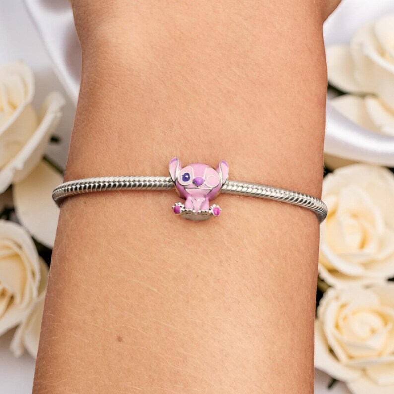 Peut inclure: Un bracelet &agrave; breloques argent&eacute; avec un charm Stitch rose et violet. Le charm repr&eacute;sente Stitch avec un &oelig;il ferm&eacute; et est orn&eacute; de d&eacute;tails roses. Le bracelet est port&eacute; sur un poignet, avec des roses blanches en arri&egrave;re-plan.