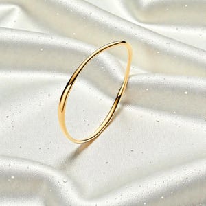 May include: A gold-colored bangle bracelet with a smooth, curved design. The bracelet is set against a backdrop of white, silky fabric, creating a luxurious and elegant aesthetic. The jewelry piece is simple yet sophisticated.
