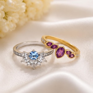 May include: A silver-toned ring featuring a central round light blue stone surrounded by clear baguette and round stones, paired with a gold-toned ring set with five marquise-cut purple stones arranged in a cluster along the band.