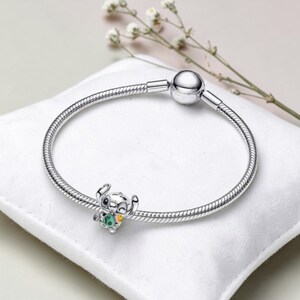 K&ouml;nnte beinhalten: Ein silbernes Charm-Armband mit einem detaillierten Charm des Charakters Stitch aus dem Disney-Film Lilo & Stitch. Der Charm ist farbenfroh und hat gr&uuml;ne, gelbe und wei&szlig;e Akzente. Das Armband liegt auf einer wei&szlig;en Oberfl&auml;che.