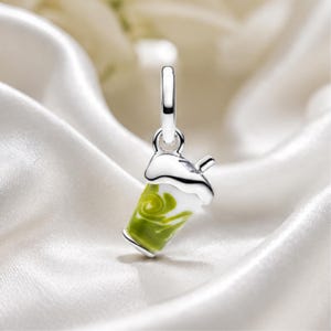 May include: A silver charm shaped like a cup of matcha latte, featuring a polished silver top with a small straw and a white and green swirled enamel body. The charm includes a circular silver bail at the top for attachment to a bracelet or necklace.