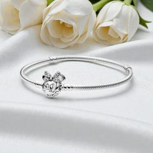 May include: A silver snake chain bracelet featuring a clasp shaped like the head of a cartoon mouse with a bow. The bow is embellished with small, clear, sparkling stones. The bracelet has a smooth, polished metallic finish and a circular shape.