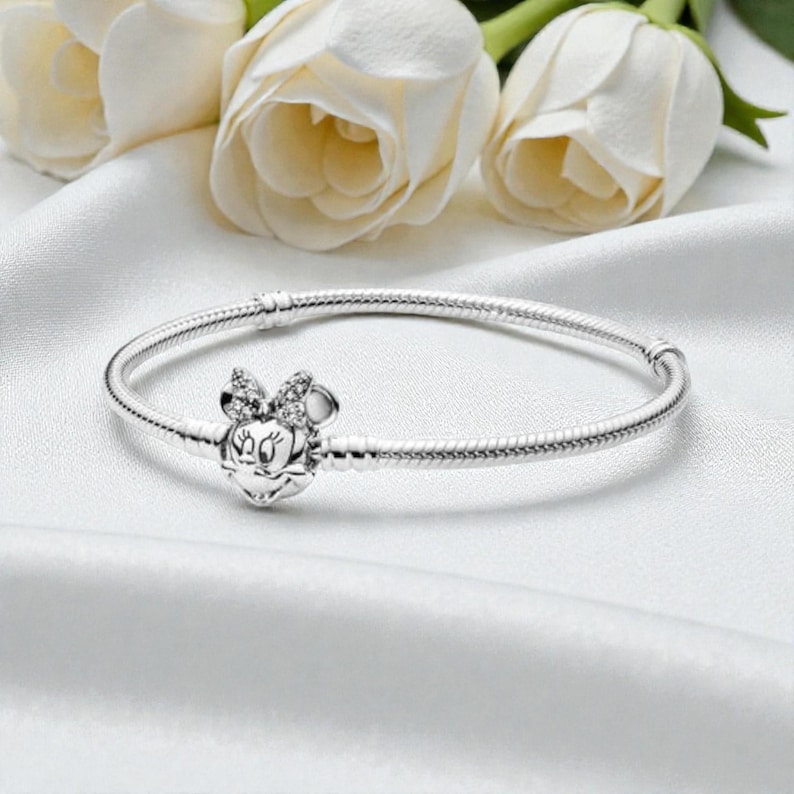 May include: A silver snake chain bracelet featuring a clasp shaped like the head of a cartoon mouse with a bow. The bow is embellished with small, clear, sparkling stones. The bracelet has a smooth, polished metallic finish and a circular shape.
