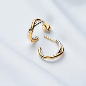 Könnte beinhalten: Ein Paar zweifarbige Creolen. Die Ohrringe haben ein verdrehtes Design mit einer goldfarbenen Außenseite und einer silberfarbenen Innenseite. Die Ohrringe werden auf einer weißen Oberfläche präsentiert.
