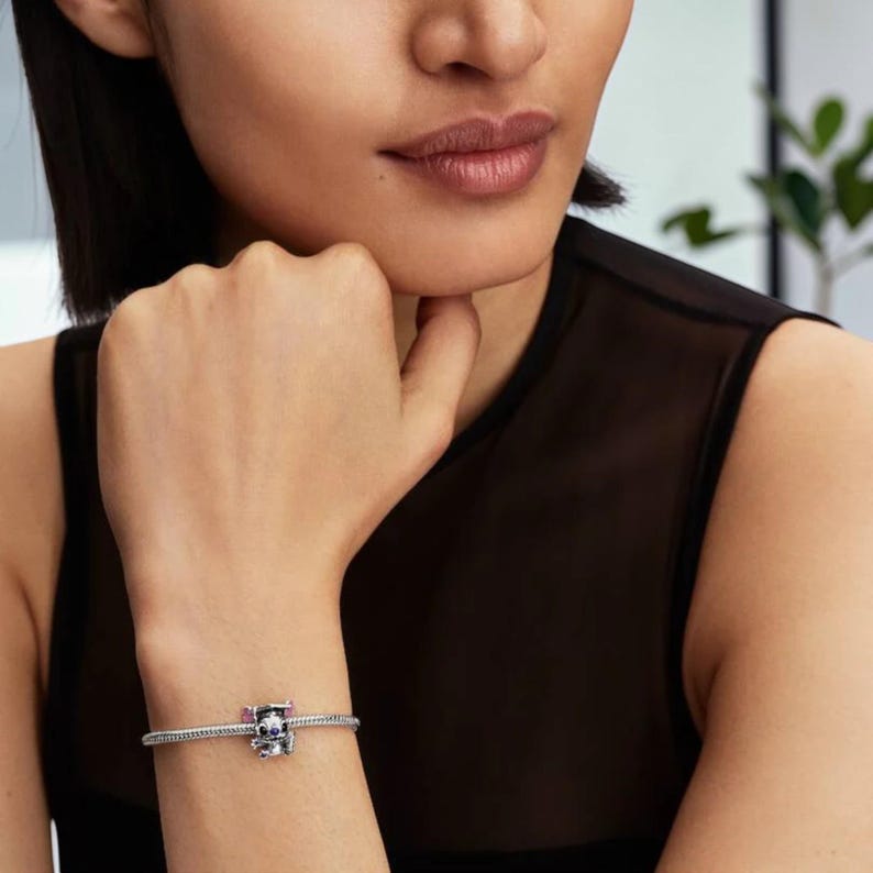 May include: A silver bracelet with a decorative charm featuring a skull design with pink and purple accents. The charm is the focal point of the bracelet, which is worn on the wrist.