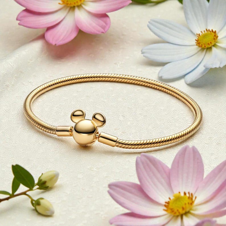 Pu&ograve; includere: Un braccialetto dorato con chiusura a forma di Mickey Mouse. Il braccialetto a catena a serpente &egrave; presentato su una superficie bianca, con fiori rosa e bianchi sullo sfondo. Il braccialetto &egrave; un gioiello.