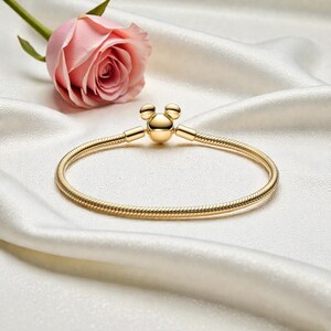 Pu&ograve; includere: Un braccialetto dorato con chiusura a forma di Mickey Mouse. Il braccialetto &egrave; realizzato con una catena flessibile a forma di serpente. Una rosa rosa &egrave; sullo sfondo, su una superficie di raso bianco. &Egrave; un gioiello.