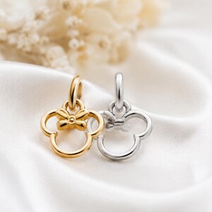 Puede incluir: Dos colgantes de metal, uno dorado y otro plateado, cada uno con forma de cabeza estilizada con orejas redondeadas y un peque&ntilde;o lazo en la parte superior. Cada colgante cuenta con una anilla circular para sujetarlo a una cadena o collar.